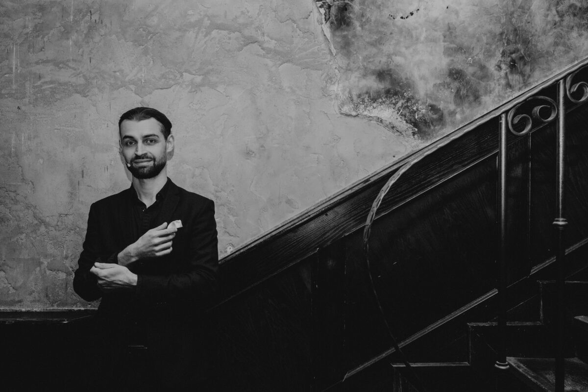 Fotograf na impreze firmowa weles bar warszawa23 Mężczyzna w ciemnym garniturze stoi obok drewnianych schodów, krzyżując ręce i lekko się uśmiechając, na tle teksturowanej, zwietrzałej ściany. Zdjęcie jest czarno-białe.