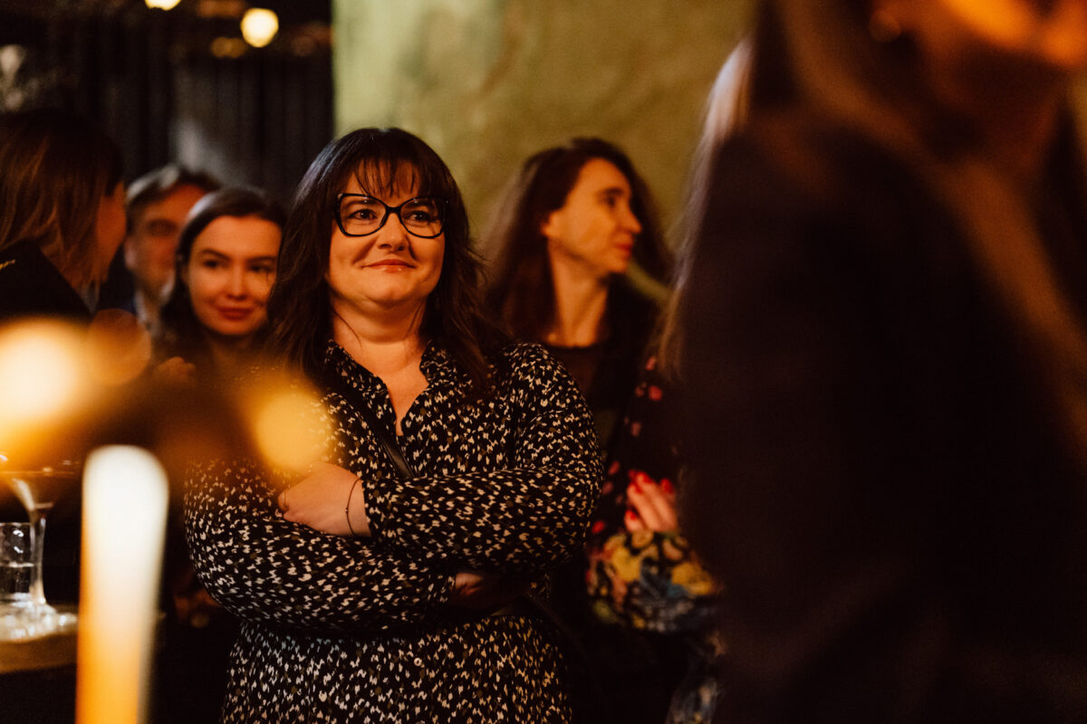 Fotograf na impreze firmowa weles bar warszawa24 1 Kobieta w okularach i ciemnych włosach stoi ze skrzyżowanymi ramionami, uśmiechając się w ciepło oświetlonym pomieszczeniu z kilkoma innymi osobami w tle, co sugeruje wydarzenie towarzyskie lub networkingowe.