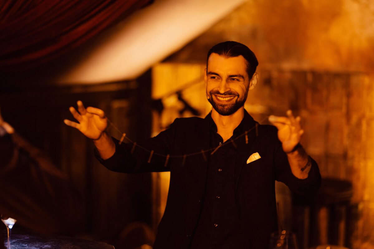 Fotograf na impreze firmowa weles bar warszawa28 Uśmiechnięty mężczyzna w ciemnej kurtce wykonuje magiczną sztuczkę, trzymając cienki, elastyczny przedmiot rozciągnięty między rękami w ciepło oświetlonym, rustykalnym wnętrzu.