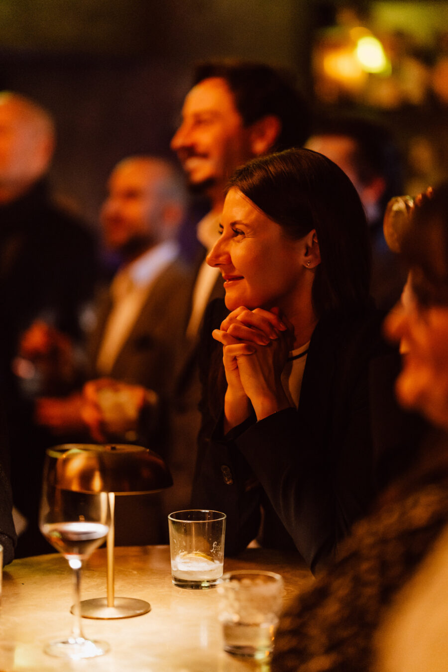 Fotograf na impreze firmowa weles bar warszawa29 Kobieta siedzi przy słabo oświetlonym barze, uśmiechając się z rękami splecionymi pod brodą. Jest otoczona przez innych ludzi w ciepłej, żywej atmosferze. Na stoliku przed nią stoją kieliszki i lampa.