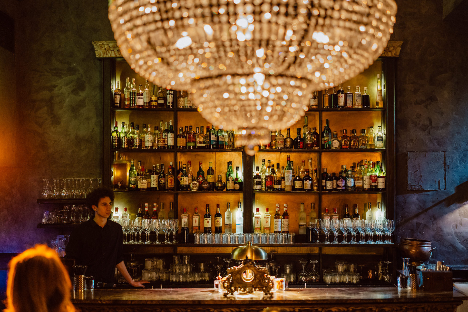Fotograf na impreze firmowa weles bar warszawa3 Słabo oświetlony bar z dużym, ozdobnym żyrandolem na pierwszym planie. Na półkach za barem stoją różne butelki z alkoholem. Barman stoi za ladą, a jedna osoba siedzi przed barem.
