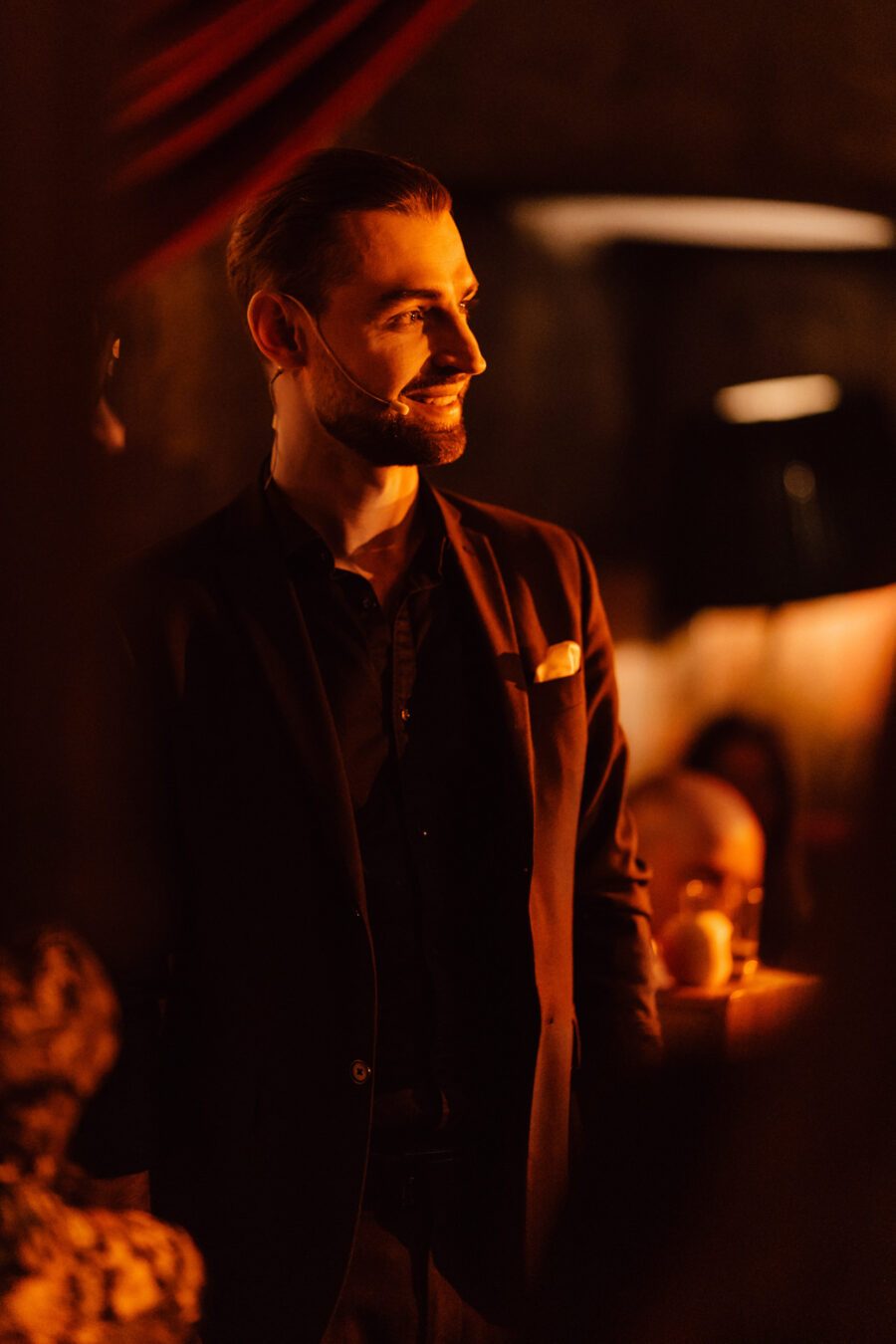 Fotograf na impreze firmowa weles bar warszawa30 Mężczyzna w ciemnym garniturze z poszetką stoi w ciepłym, dramatycznym oświetleniu, nosząc mikrofon słuchawkowy i lekko się uśmiechając, z rozmytą publicznością i przyciemnionym tłem za nim.