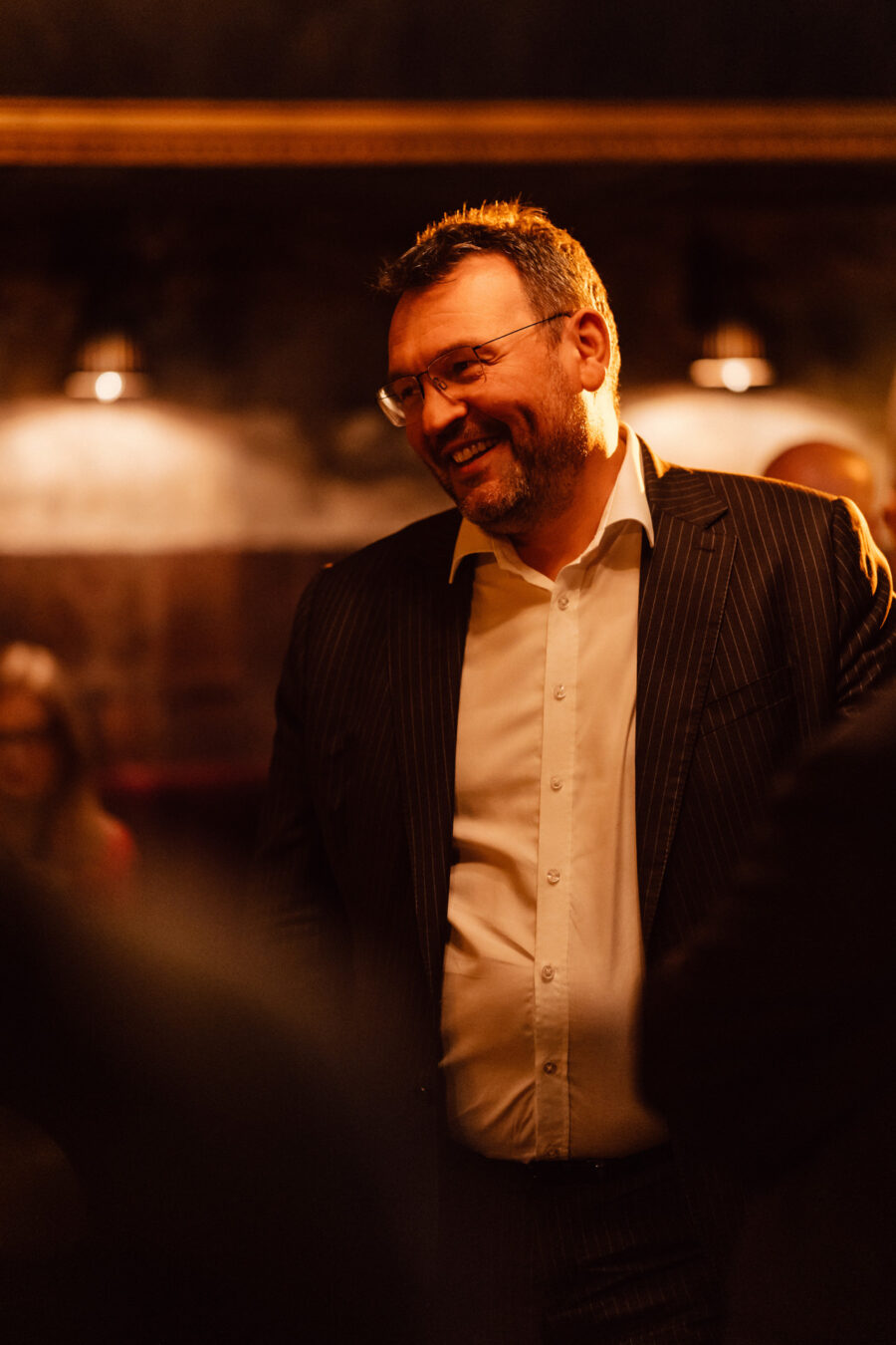 Fotograf na impreze firmowa weles bar warszawa31 Mężczyzna w prążkowanym garniturze i okularach uśmiecha się ciepło w słabo oświetlonym pokoju z miękkim, ciepłym oświetleniem i rozmytymi postaciami w tle.