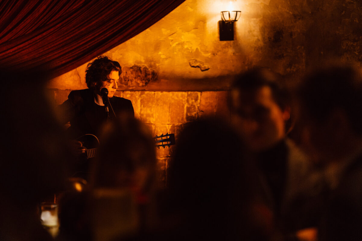 Fotograf na impreze firmowa weles bar warszawa34 Muzyk stoi przy ciepłym, słabym oświetleniu, grając na gitarze i śpiewając do mikrofonu w kameralnym, rustykalnym miejscu z ludźmi na widowni.