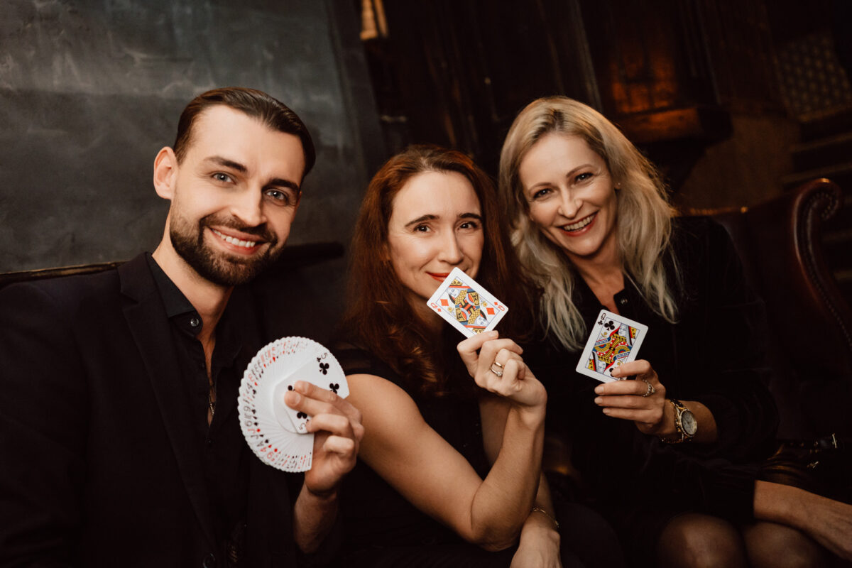 Fotograf na impreze firmowa weles bar warszawa35 Trzy osoby siedzące razem w pomieszczeniu, uśmiechnięte i trzymające karty do gry. Mężczyzna po lewej stronie trzyma wachlarzową talię, podczas gdy dwie kobiety obok niego pokazują zakryte karty, wydając się cieszyć magiczną sztuczką lub grą karcianą.
