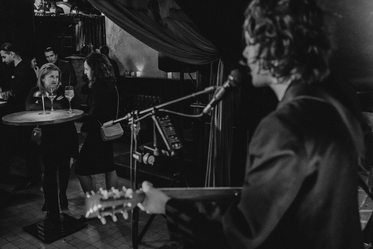 Fotograf na impreze firmowa weles bar warszawa36 Muzyk występuje z gitarą i mikrofonem na scenie, podczas gdy dwie kobiety stoją w pobliżu małego okrągłego stolika z napojami, uważnie obserwując w słabo oświetlonym, intymnym miejscu.