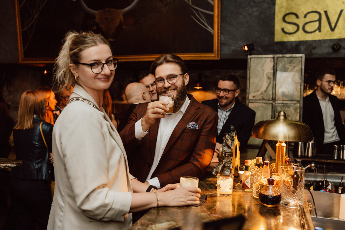 Fotograf na impreze firmowa weles bar warszawa37 1 Kobieta i mężczyzna stoją razem przy barze, uśmiechając się i trzymając drinki. Inni ludzie spotykają się w tle, a scena jest ciepło oświetlona ze stylową, przytulną atmosferą.
