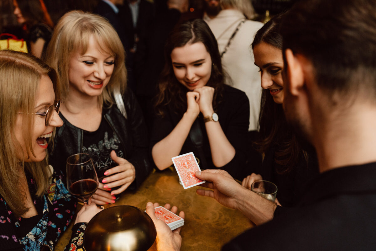 Fotograf na impreze firmowa weles bar warszawa39 Grupa kobiet siedzi przy stole, uśmiechając się i obserwując magika wykonującego sztuczkę karcianą. Jedna z kobiet trzyma kieliszek wina, podczas gdy inne wyglądają na zaintrygowane i rozbawione. Otoczenie wydaje się żywe i towarzyskie.