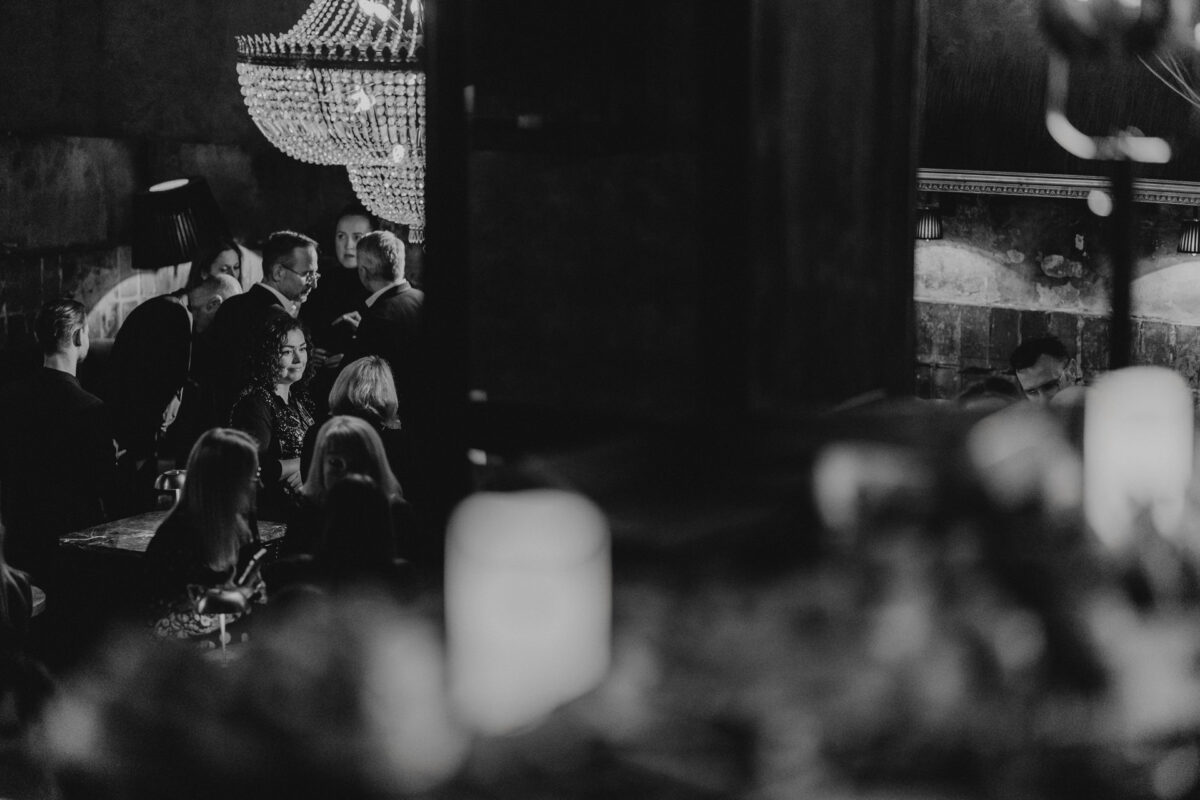 Fotograf na impreze firmowa weles bar warszawa40 Czarno-białe zdjęcie przedstawiające ludzi w słabo oświetlonym pokoju z dużym żyrandolem nad głową, niektórzy siedzą, a niektórzy stoją, tworząc intymną i elegancką atmosferę.