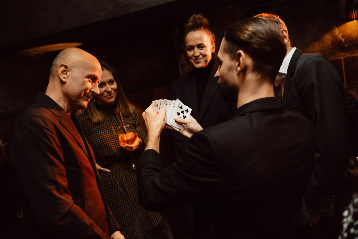 Fotograf na impreze firmowa weles bar warszawa41 Grupa ludzi w formalnych strojach obserwuje mężczyznę wykonującego sztuczkę karcianą, trzymając rozłożoną talię kart. Widzowie wydają się zaangażowani i uśmiechnięci w słabo oświetlonym pomieszczeniu.