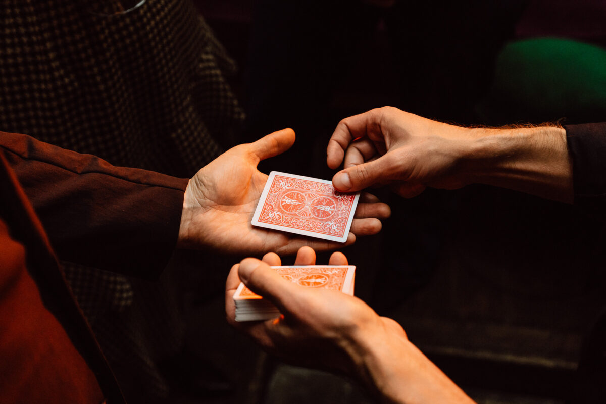 Fotograf na impreze firmowa weles bar warszawa42 Zbliżenie rąk trzech osób trzymających i podających talie kart do gry, sugerujące grę karcianą lub magiczną sztuczkę, na ciemnym tle.
