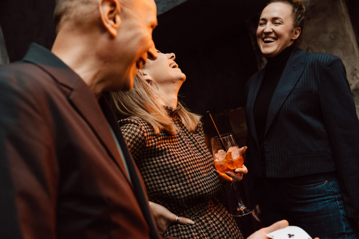 Fotograf na impreze firmowa weles bar warszawa43 Trzy osoby stoją razem w pomieszczeniu, śmiejąc się i uśmiechając. Jedna kobieta trzyma koktajl z lodem i słomką, podczas gdy druga osoba trzyma kartę do gry. Atmosfera wydaje się żywa i radosna.