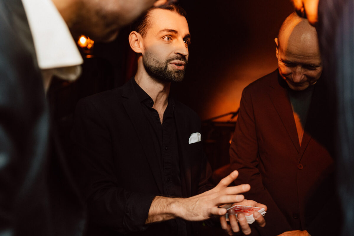 Fotograf na impreze firmowa weles bar warszawa44 Mężczyzna w ciemnym garniturze trzyma karty do gry i wydaje się wykonywać sztuczkę karcianą dla małej grupy ludzi, którzy uważnie obserwują w słabo oświetlonym pokoju.