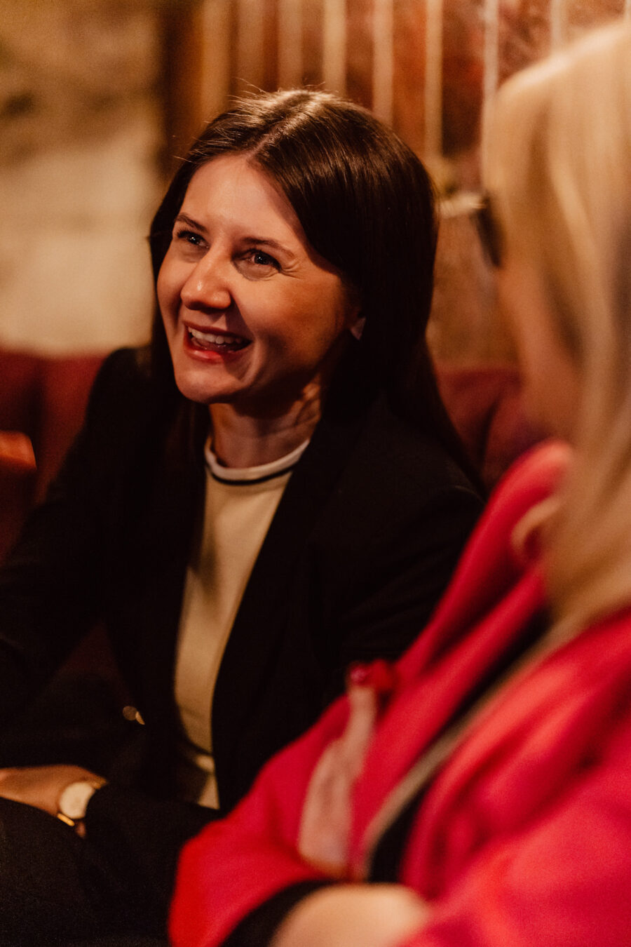 Fotograf na impreze firmowa weles bar warszawa45 1 Dwie kobiety siedzą razem w pomieszczeniu i rozmawiają. Jedna kobieta o ciemnych włosach uśmiecha się szeroko, podczas gdy druga, o blond włosach i ubrana w jasną kurtkę, jest widziana z boku. Otoczenie wydaje się ciepłe i przytulne.