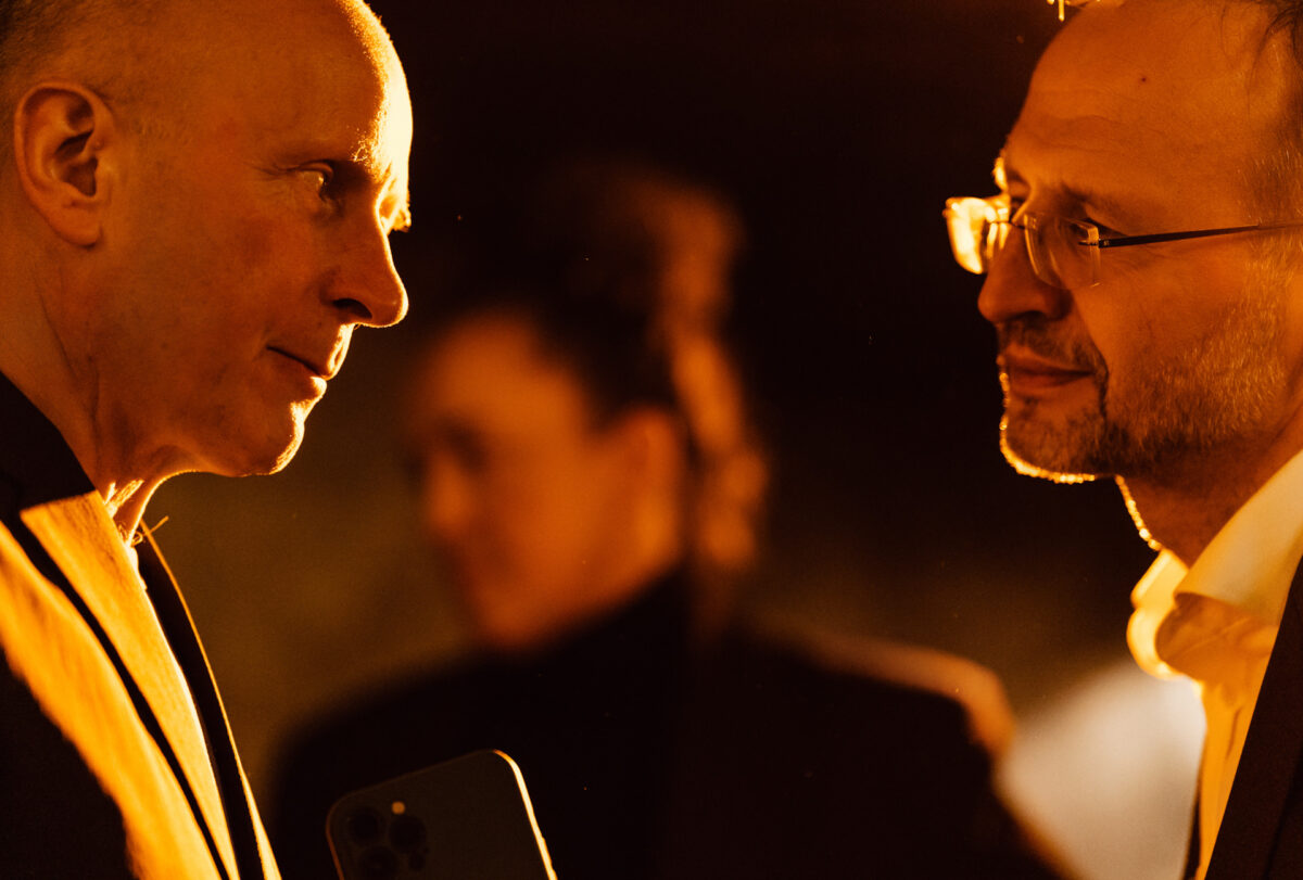 Fotograf na impreze firmowa weles bar warszawa47 Dwaj mężczyźni w garniturach stoją naprzeciw siebie w rozmowie, ciepło oświetleni pomarańczowymi tonami. Kobieta stoi w rozmytym tle, tworząc poczucie głębi i skupiając się na interakcji mężczyzn.