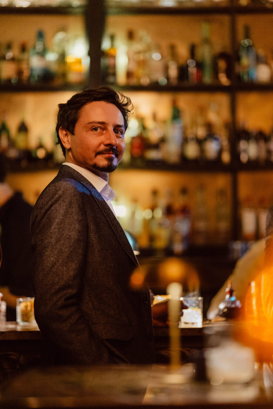 Fotograf na impreze firmowa weles bar warszawa48 Mężczyzna w garniturze stoi przy słabo oświetlonym barze, spoglądając przez ramię i uśmiechając się. Półki wypełnione butelkami są rozmyte w tle, tworząc ciepłą, zachęcającą atmosferę.