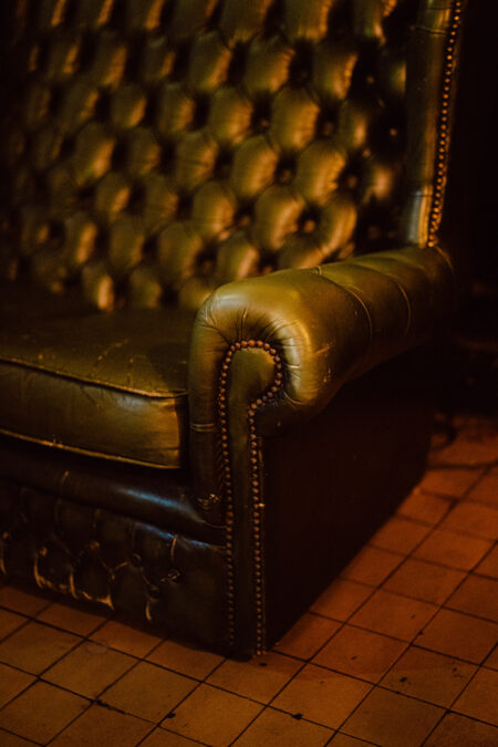 Fotograf na impreze firmowa weles bar warszawa5 Zbliżenie na zabytkowy, ciemnozielony skórzany fotel z tapicerką obitą guzikami i ozdobnymi mosiężnymi gwoździami, umieszczony na brązowej podłodze wyłożonej kafelkami w ciepłym oświetleniu.