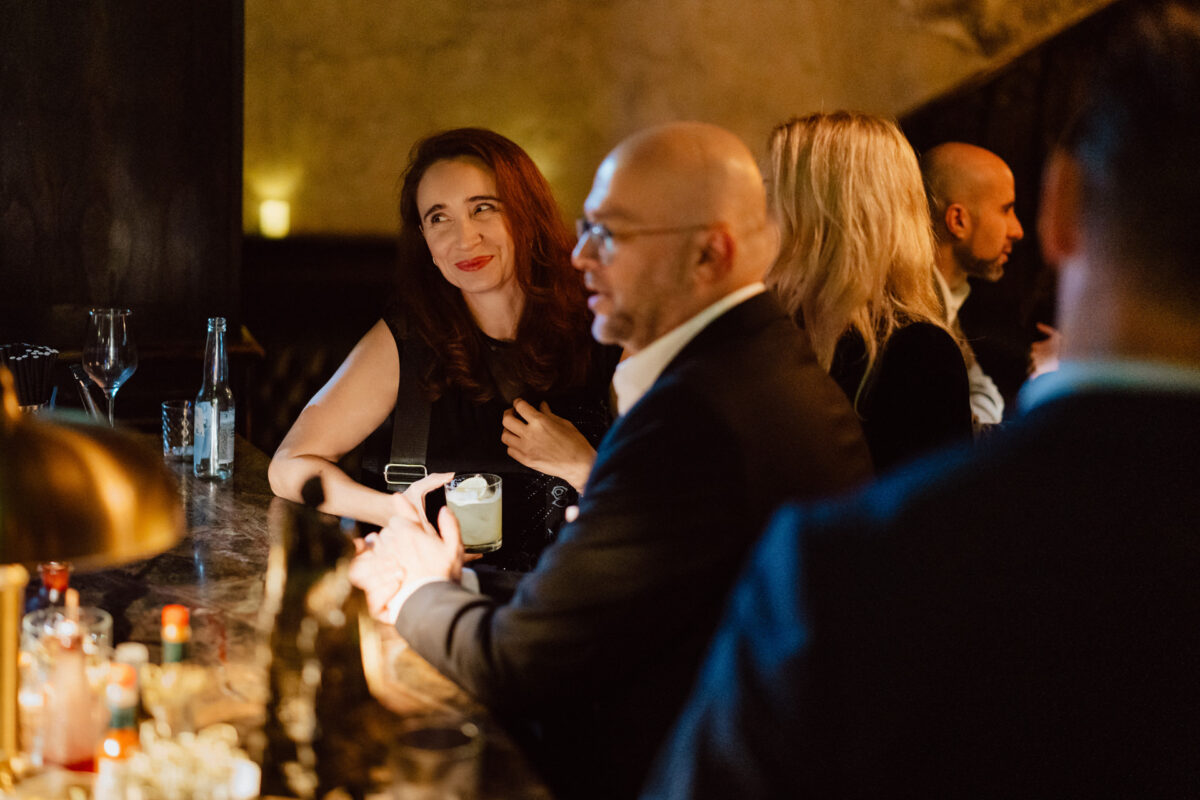 Fotograf na impreze firmowa weles bar warszawa50 1 Kobieta z długimi rudymi włosami uśmiecha się, trzymając drinka w słabo oświetlonym barze, w otoczeniu innych osób zaangażowanych w rozmowę. Butelki i szklanki stoją na ladzie barowej.