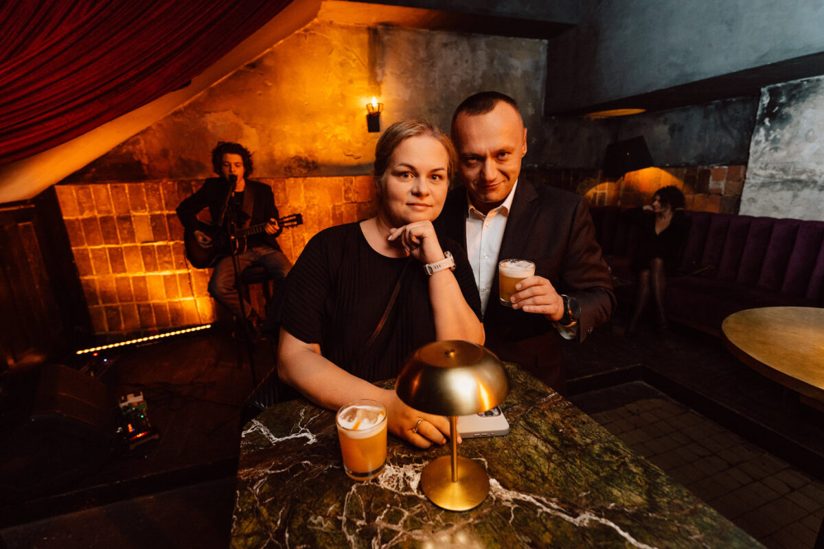Fotograf na impreze firmowa weles bar warszawa51 1 Kobieta i mężczyzna stoją razem przy marmurowym stoliku barowym, każde trzyma drinka, uśmiechając się do kamery w słabo oświetlonym, przytulnym barze. W tle muzyk gra na gitarze, dodając do relaksującej atmosfery.