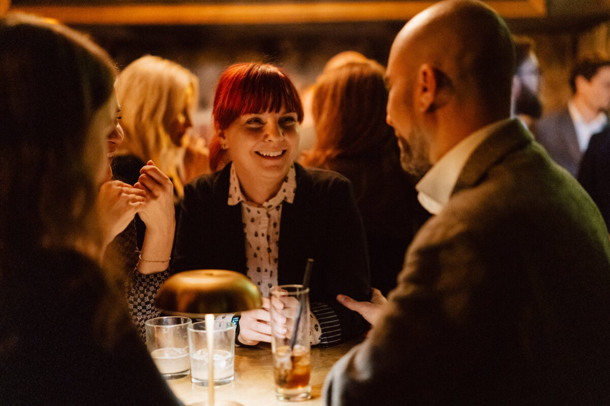 Fotograf na impreze firmowa weles bar warszawa52 1 Grupa ludzi siedzi przy słabo oświetlonym stoliku w barze, uśmiechając się i rozmawiając. Na stole stoją napoje, a atmosfera jest ciepła i ożywiona.
