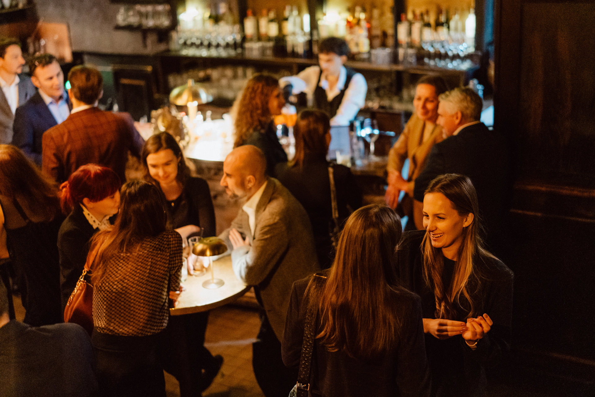 Słabo oświetlony bar z dużym, ozdobnym żyrandolem na pierwszym planie. Na półkach za barem stoją różne butelki z alkoholem. Barman stoi za ladą, a jedna osoba siedzi przed barem.