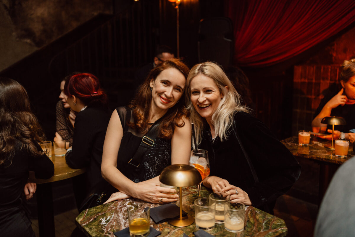 Fotograf na impreze firmowa weles bar warszawa54 Dwie kobiety siedzą blisko siebie i uśmiechają się do kamery w słabo oświetlonym barze lub salonie, z napojami na marmurowym stole przed nimi i innymi osobami rozmawiającymi w tle.
