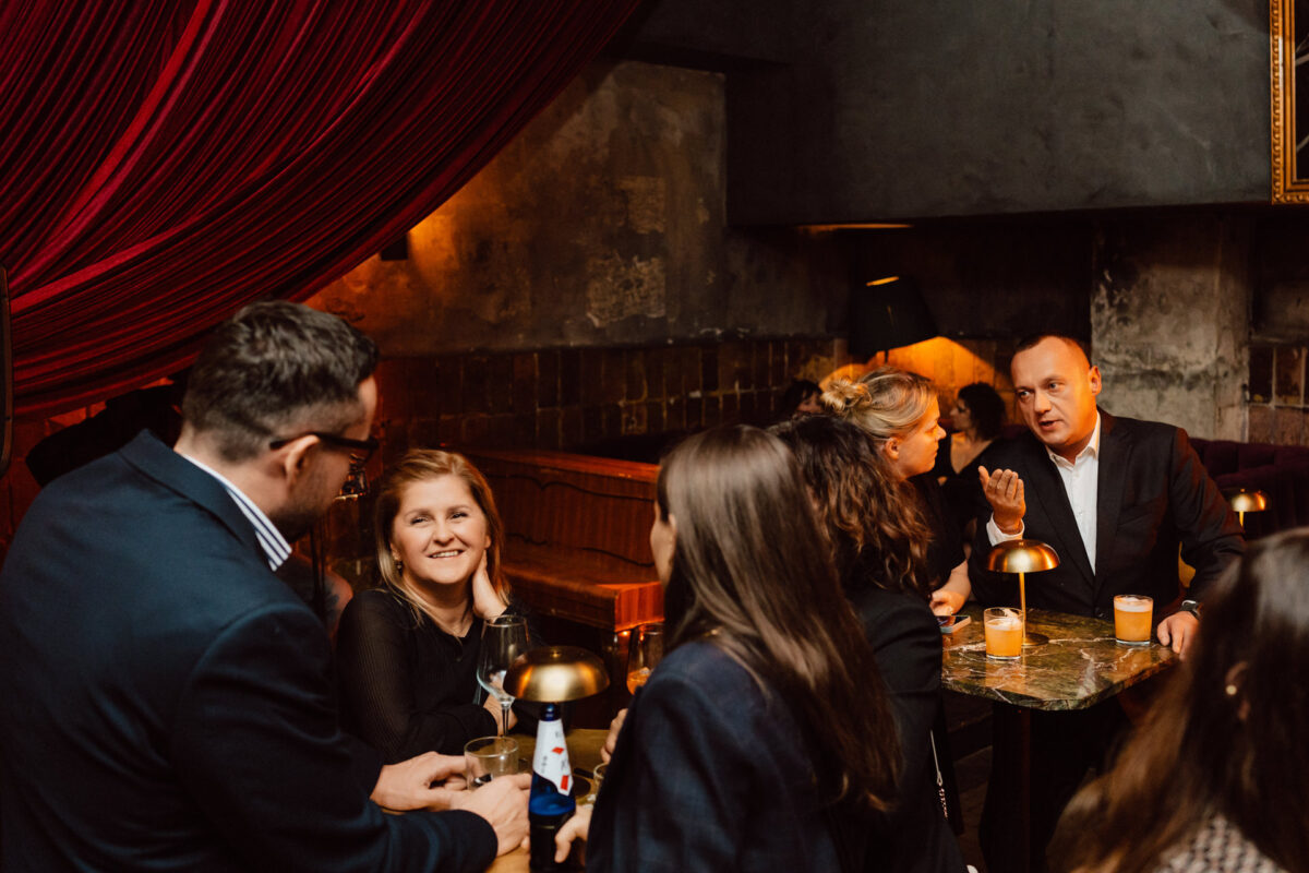 Fotograf na impreze firmowa weles bar warszawa55 1 Grupa ludzi w formalnych strojach spotyka się przy stolikach z drinkami w słabo oświetlonym barze. Jeden z mężczyzn gestykuluje, podczas gdy inni uśmiechają się i rozmawiają przy ciepłym oświetleniu i czerwonej draperii.