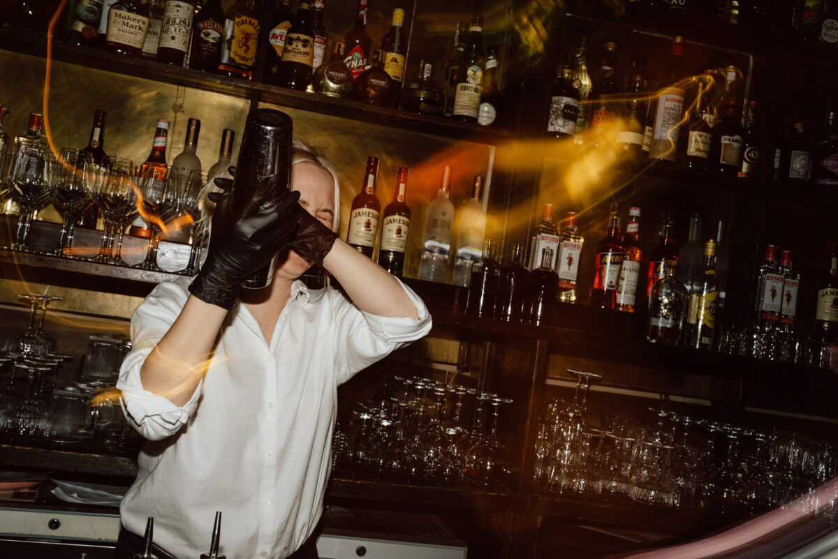 Fotograf na impreze firmowa weles bar warszawa56 Barman w białej koszuli i czarnych rękawiczkach potrząsa shakerem do koktajli za słabo oświetlonym barem, z półkami pełnymi różnych butelek i szklanek w tle. Smugi światła tworzą dynamiczną atmosferę.