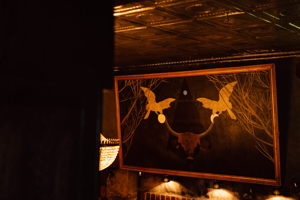 Fotograf na impreze firmowa weles bar warszawa57 Oprawiona grafika przedstawiająca dwa złote ptaki zwrócone do siebie nad rogami bawołu na ciemnym tle wisi na ścianie z misternymi płytami sufitowymi powyżej. Ciepłe oświetlenie podkreśla grafikę.