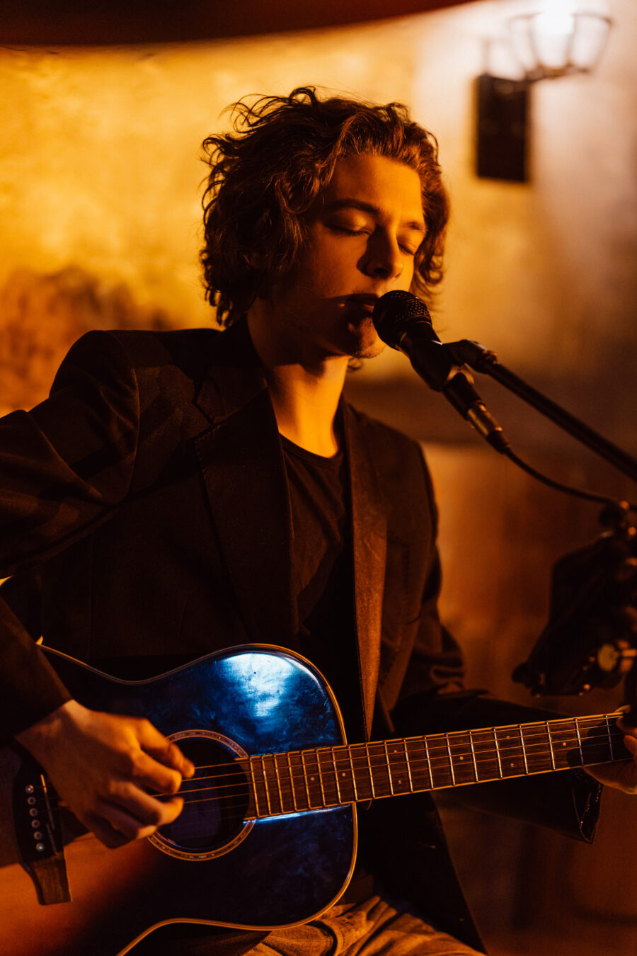 Fotograf na impreze firmowa weles bar warszawa7 Młoda osoba z falującymi włosami gra na gitarze akustycznej i śpiewa do mikrofonu na ciepło oświetlonej scenie, ubrana w ciemną marynarkę.