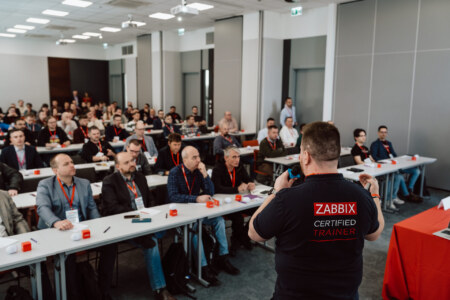 Fotorelacja z konferencji - Mężczyzna ubrany w koszulkę "Zabbix Certified Trainer" przemawia do dużej publiczności siedzącej przy stołach w sali konferencyjnej, której uczestnicy uważnie słuchają, a niektórzy robią notatki.