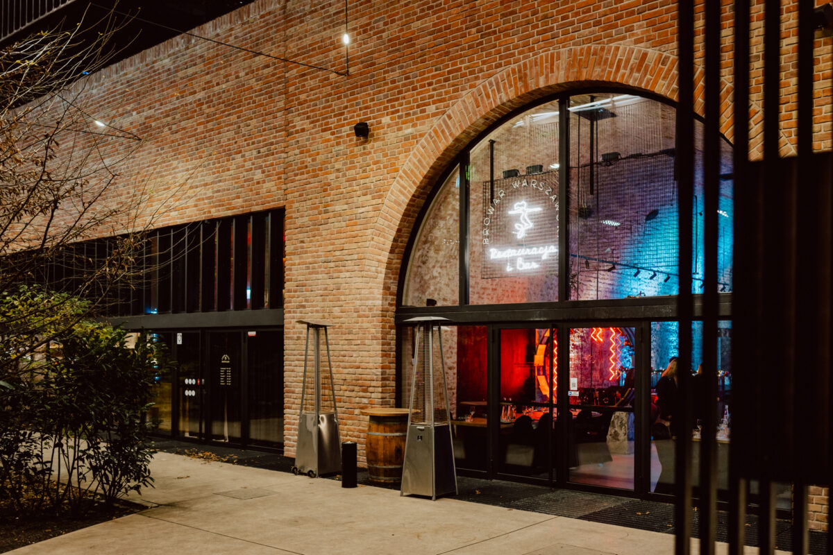 Ceglany budynek z dużymi łukowymi oknami i szklanymi drzwiami, ciepło oświetlony wewnątrz. Nad wejściem świeci neon z tańczącą postacią, a na zewnątrz przy drzwiach stoi beczka. Scena wieczorna.