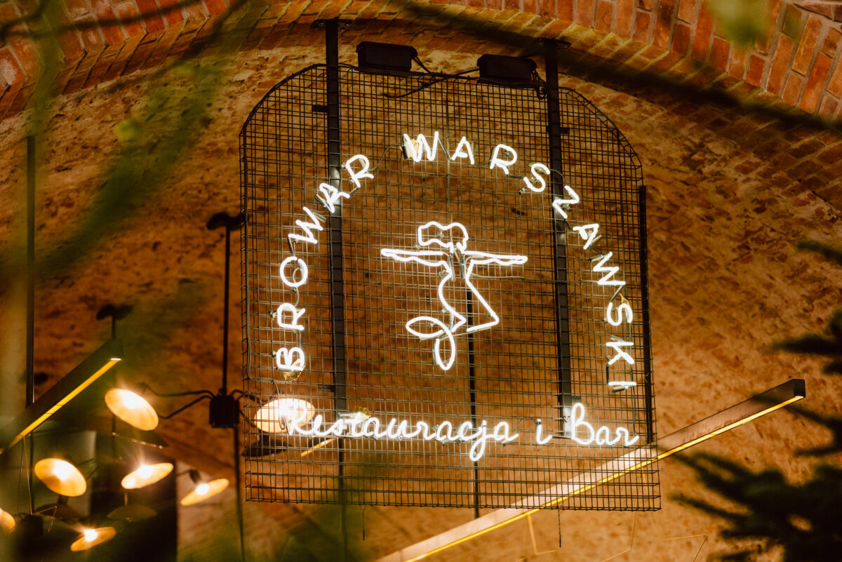 Świecący neon z napisem "Browar Warszawski Restauracja i Bar" z ilustracją kobiety trzymającej dwa kufle piwa, zamontowany na ceglanym łukowym suficie.