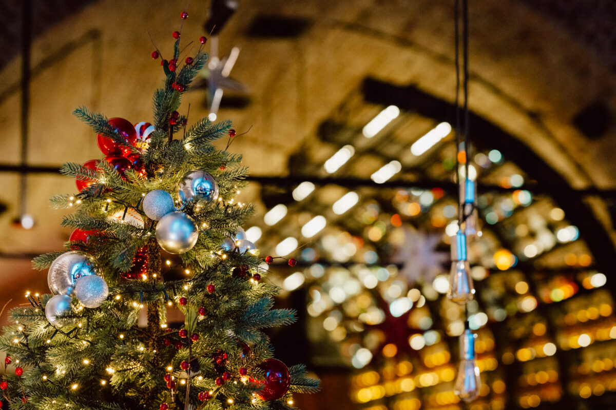 Udekorowana choinka z czerwonymi i srebrnymi ozdobami i światełkami stoi w pomieszczeniu, z rozmytym tłem łukowatych okien i świecących światełek.