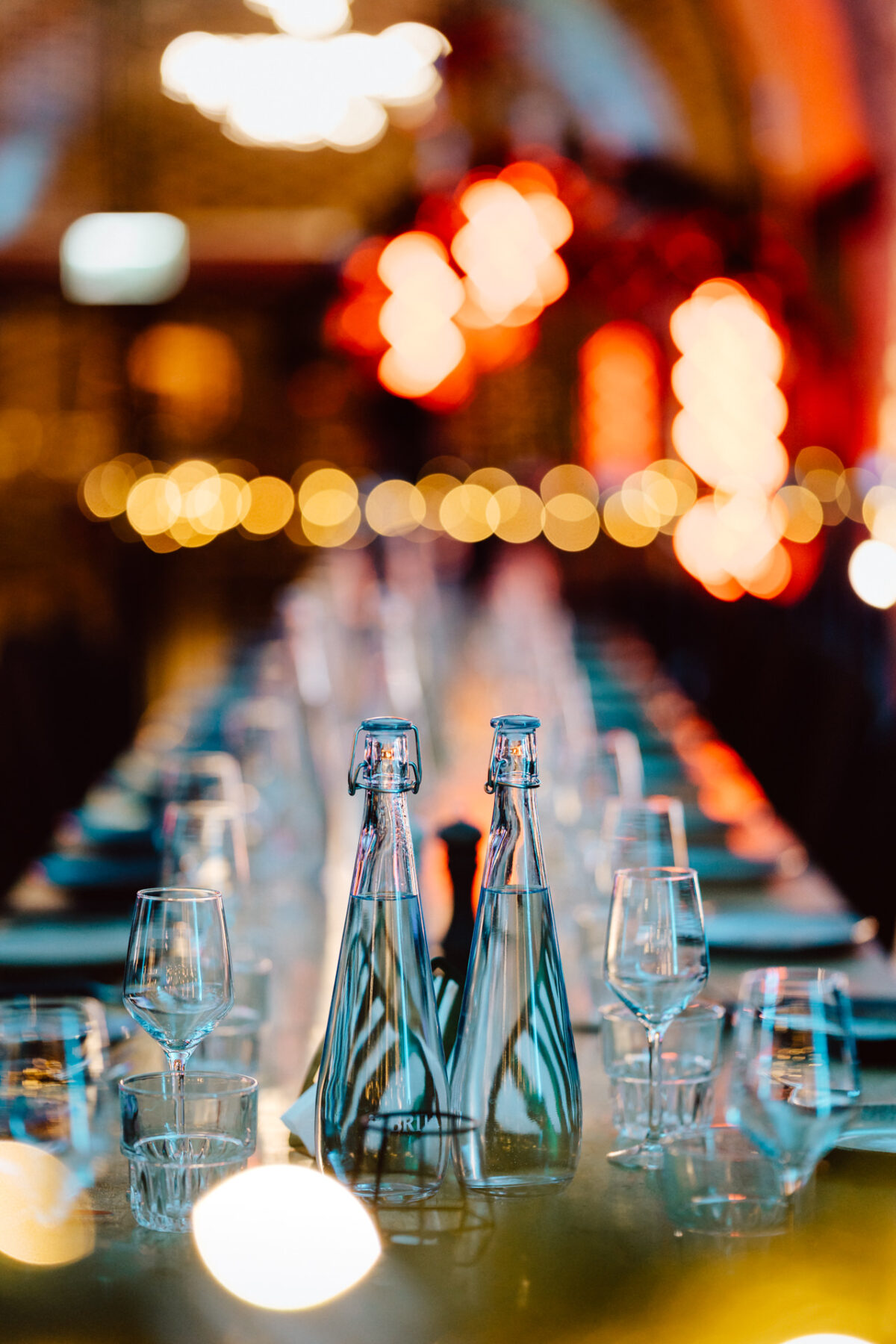 Dwie szklane butelki wody i kilka pustych kieliszków do wina są starannie ułożone na odblaskowym stole jadalnym, a ciepłe, rozmyte światła tworzą świąteczną atmosferę w tle.