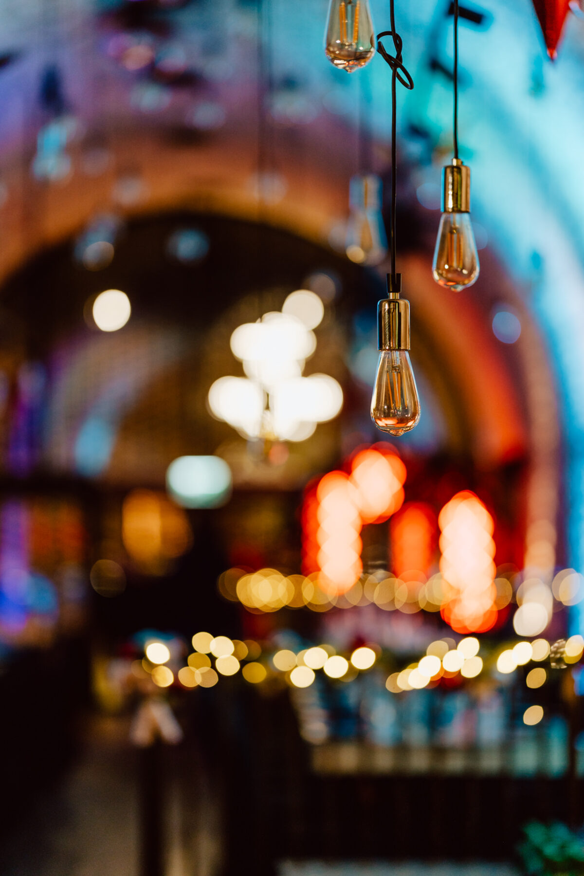 Zbliżenie na kilka wiszących żarówek w ostrości, z rozmytym tłem kolorowych świateł i żyrandolem w ciepło oświetlonej przestrzeni wewnętrznej, tworząc przytulną i świąteczną atmosferę.