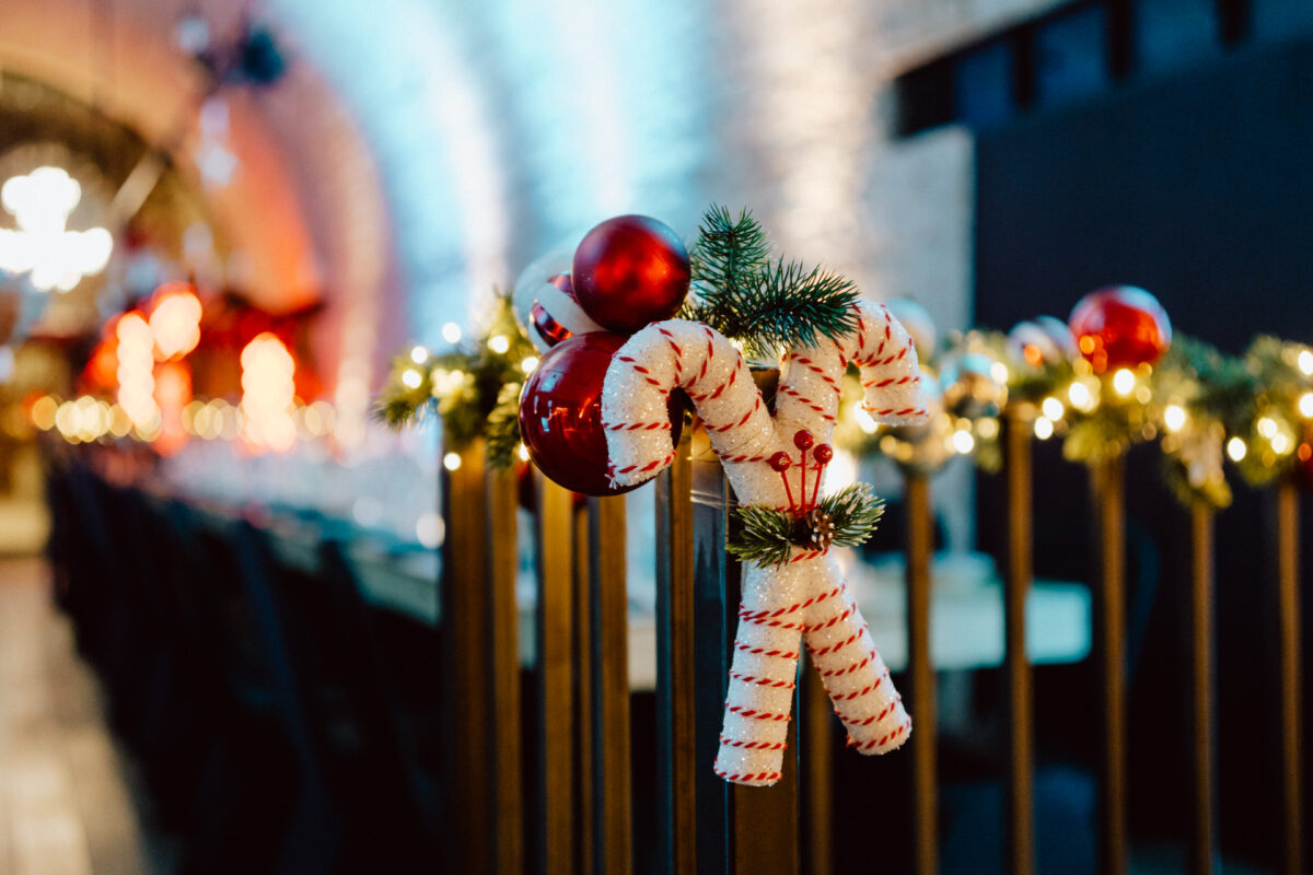 Zbliżenie na świąteczną dekorację z materiałowymi cukierkami, czerwonymi bombkami i gałązkami sosny przymocowanymi do poręczy, z rozmytymi światłami i długim, udekorowanym stołem w tle.