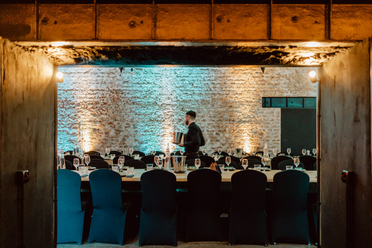 Długi stół z ciemnymi krzesłami jest ustawiony na formalne wydarzenie w rustykalnym pokoju z ceglanymi ścianami i ciepłym oświetleniem. Serwer niosący wiadro przechodzi obok stołu.