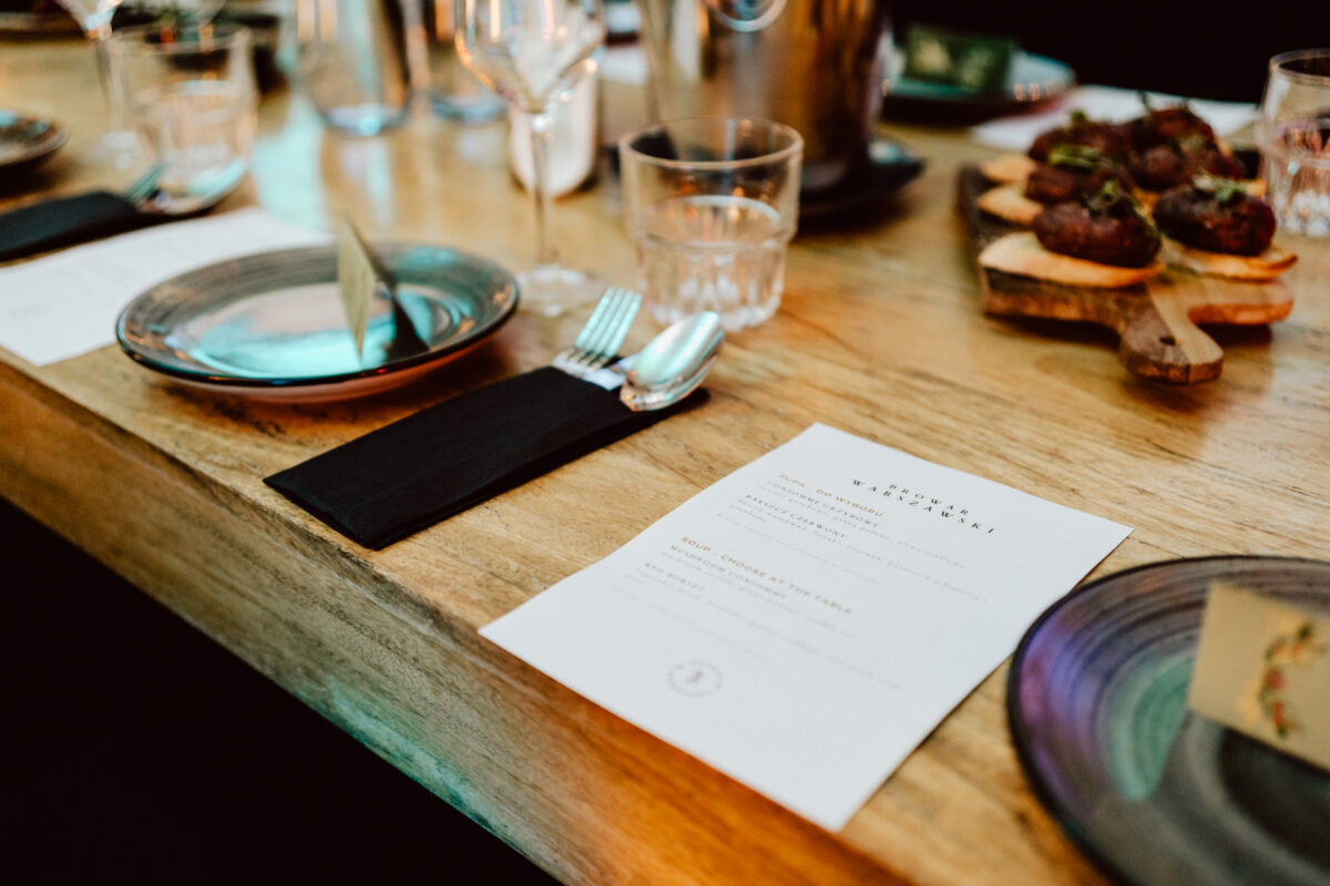 Drewniany stół nakryty do posiłku z talerzami, szklankami, przyborami na czarnych serwetkach, wydrukowanym menu i deską do serwowania z przystawkami w tle.