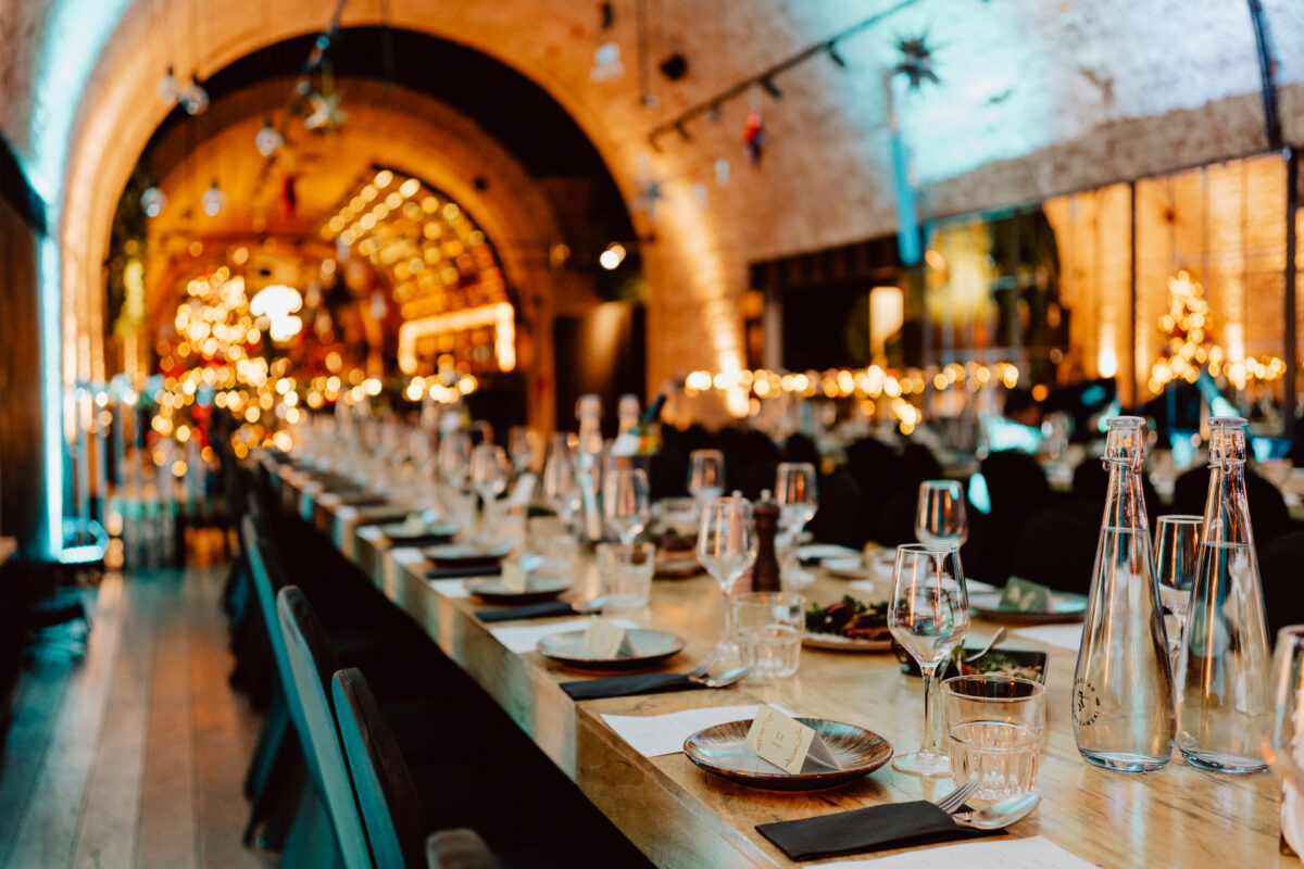 Długi stół bankietowy jest zastawiony kieliszkami, talerzami i kartami miejsc w eleganckim, ciepło oświetlonym miejscu z łukowymi sufitami i świątecznymi dekoracjami.