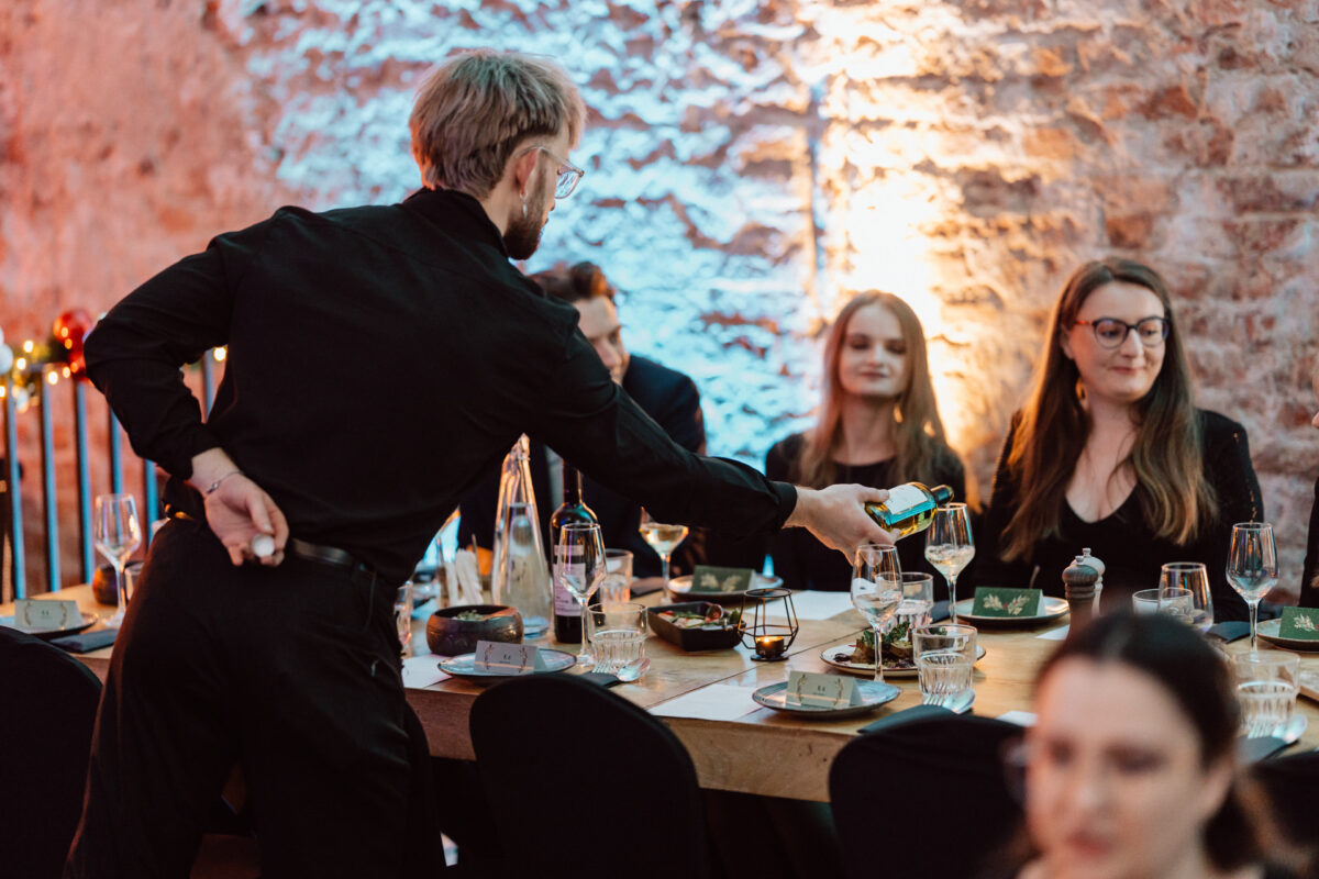 Kelner w czarnym stroju nalewa drinka gościom siedzącym przy udekorowanym stole w ciepło oświetlonej restauracji z odsłoniętymi ceglanymi ścianami. Widoczne są cztery osoby zaangażowane w rozmowę i spożywające posiłek.