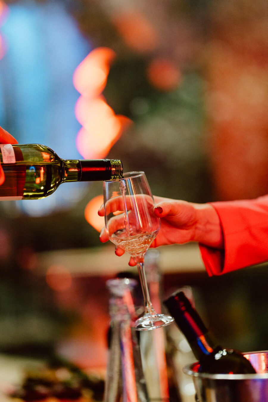 Osoba nalewa białe wino do kieliszka trzymanego przez kogoś w czerwonym rękawie, z rozmytym, kolorowym tłem i butelkami wina widocznymi na stole.