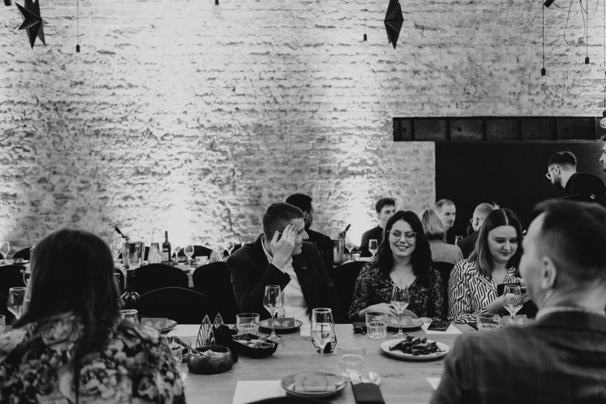 Grupa ludzi siedzi przy stole podczas oficjalnego wydarzenia w pomieszczeniu, jedząc, pijąc i rozmawiając. Kamienne ściany i delikatne oświetlenie tworzą ciepłą i elegancką atmosferę.