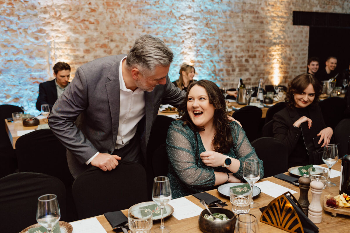 Mężczyzna w szarym garniturze pochyla się i rozmawia z uśmiechniętą kobietą siedzącą przy oficjalnym stole obiadowym, otoczonym talerzami i szklankami, z innymi gośćmi widocznymi w tle.