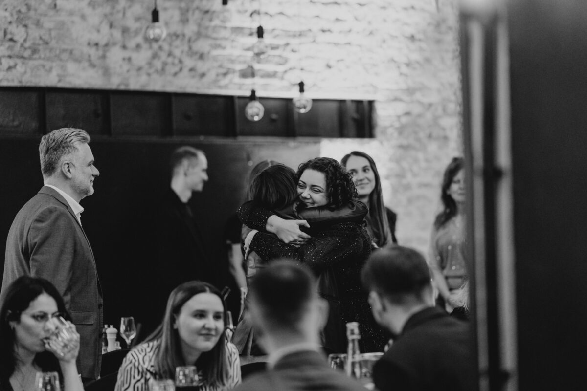 Czarno-białe zdjęcie ludzi na spotkaniu towarzyskim. W centrum dwie kobiety radośnie obejmują się, podczas gdy inni siedzą lub stoją wokół nich, rozmawiając i obserwując. W tle widać odsłoniętą cegłę i wiszące światła.