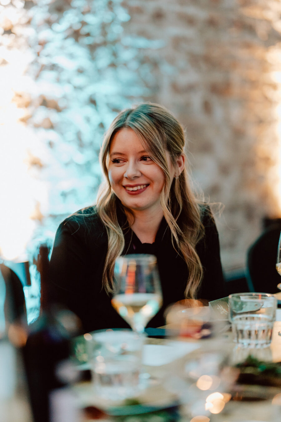 Kobieta z długimi blond włosami siedzi przy stole, uśmiechnięta, z kieliszkami do wina i talerzami przed sobą. Tło jest delikatnie oświetlone niebieskimi i żółtymi odcieniami, tworząc ciepłą, świąteczną atmosferę.