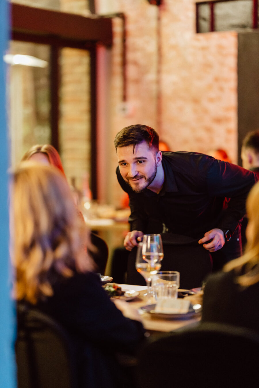Mężczyzna w czarnej koszuli pochyla się do przodu, uśmiechając się i przemawiając do grupy osób siedzących przy stoliku w restauracji zastawionym szklankami i talerzami. Atmosfera jest ciepła i zachęcająca.