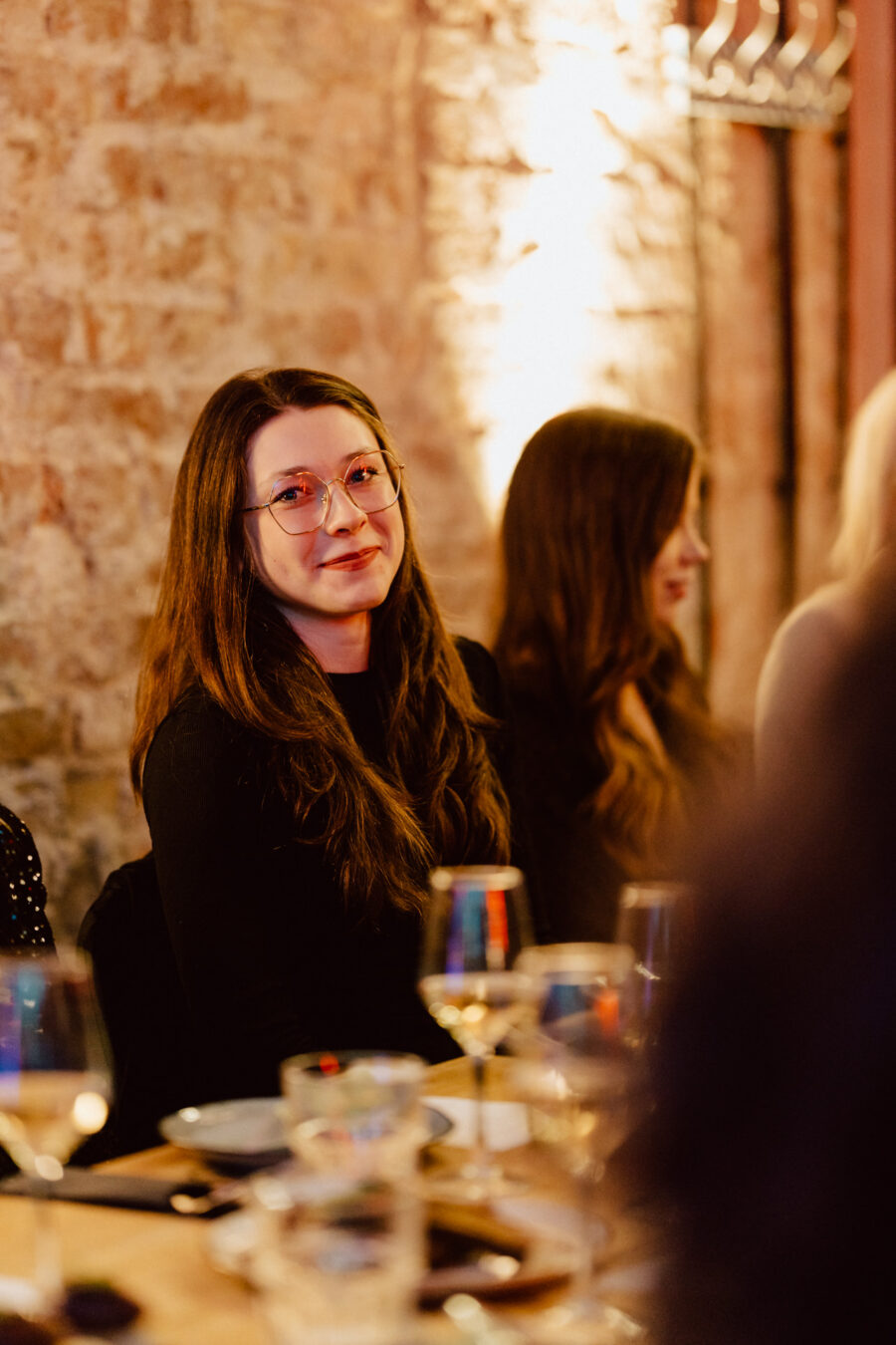 Kobieta z długimi brązowymi włosami i okularami, ubrana w czarny top, siedzi przy stole z kieliszkami do wina i talerzami, uśmiechając się lekko w ciepło oświetlonej restauracji z odsłoniętymi ceglanymi ścianami.