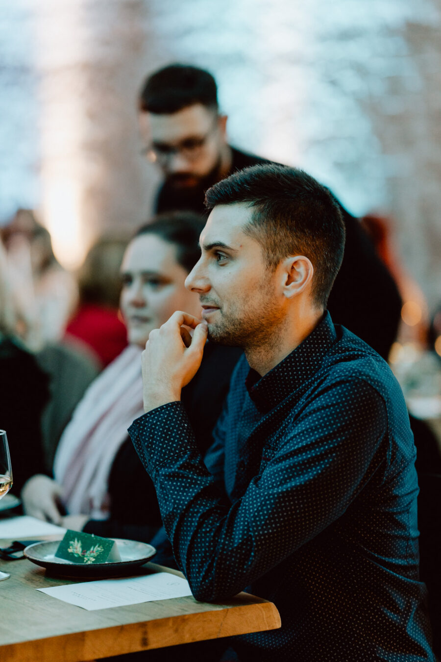 Mężczyzna siedzi przy stole w słabo oświetlonym pokoju, opierając podbródek na dłoni i patrząc przed siebie w zamyśleniu. Wokół niego siedzą inni ludzie, a na stole stoi kieliszek do wina i talerz.