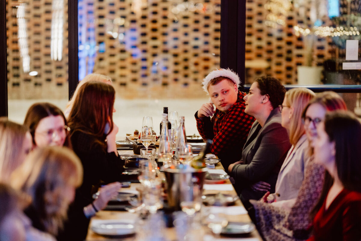 Grupa ludzi siedzi przy długim stole w ciepło oświetlonej restauracji. Jedna osoba w centrum ma na sobie czapkę Mikołaja i czerwoną koszulę w kratę, patrząc na kogoś w pobliżu, podczas gdy inni rozmawiają i cieszą się posiłkiem.